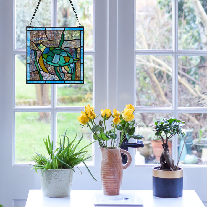 10.5"H Tommy the Green Sea Turtle Stained Glass Window Panel
 hanging on a window with potted plants and flowers on a table.