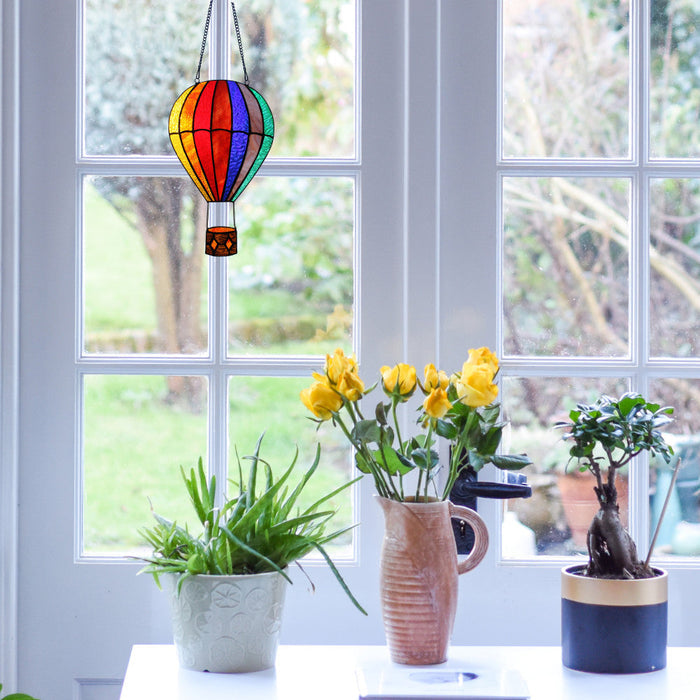 11"H Otis Red Hot Air Balloon Stained Glass Window Panel hanging above a table with potted plants and a vase of yellow flowers.