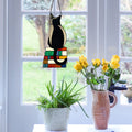 12.5"H Kate Black Library Cat Stained Glass Window Panel hanging on a window with a vase of yellow flowers and a plant below.