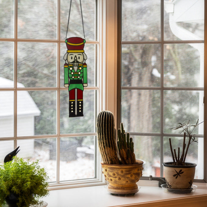 14"H Hans the Holiday Nutcracker Stained Glass Window Panel hanging in a window with potted plants below