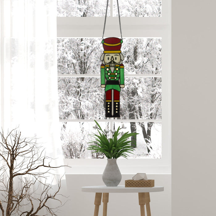 14"H Hans the Holiday Nutcracker Stained Glass Window Panel hanging by a window with a snowy outdoor view