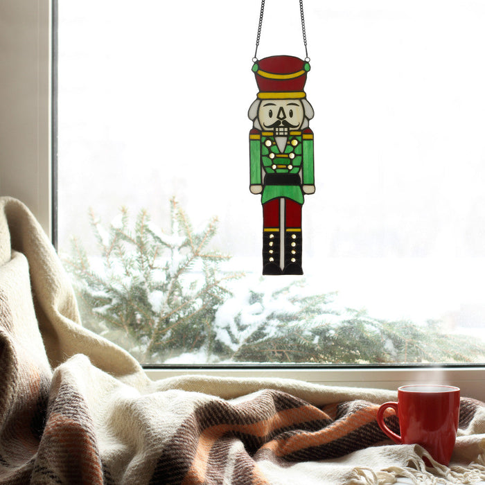 14"H Hans the Holiday Nutcracker Stained Glass Window Panel hanging in a window with a red mug on a blanket.