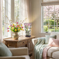 15.5" H Yara Multi Colored Stained Glass Vase-Shaped Table Lamp in a living room with a wooden table and floral arrangement, near a window with cherry blossoms outside.