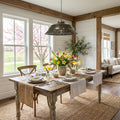 16" W Delano Antique Bronze Metal Pendant Light in a dining room with a wooden table set for a meal, flowers, and a view of cherry blossoms outside.