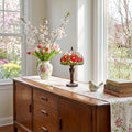 17" H Genevieve Red Flowers Stained Glass Candlestick Table Lamp in a wooden sideboard with floral arrangement, and books in a bright room with windows.