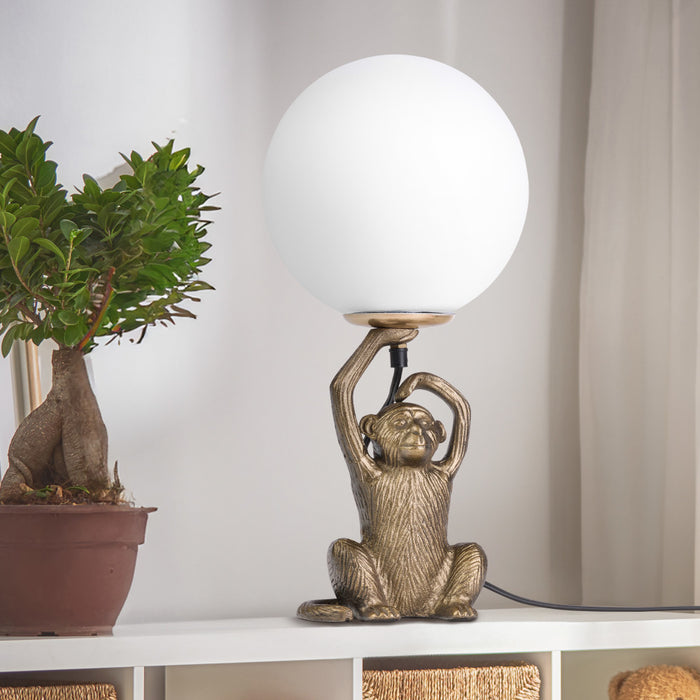 17" H Gold Finish Monkey Table Lamp with Glass Globe Shade next to a potted plant on a shelf.