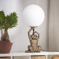 17" H Gold Finish Monkey Table Lamp with Glass Globe Shade next to a potted plant on a shelf.