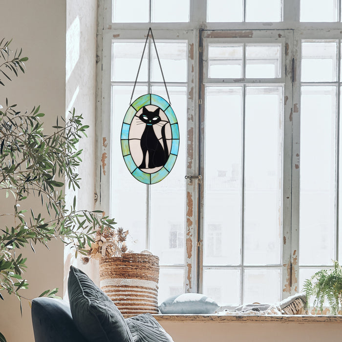 17" H Tiffany-Style Halloween Cat Stained Glass Window Panel hangs from a black cord in a bright, sunlit room with a large white window, potted plants, and a woven basket on the windowsill.