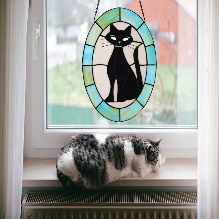17" H Tiffany-Style Halloween Cat Stained Glass Window Panel hanging on a window above a cat lying on a windowsill.