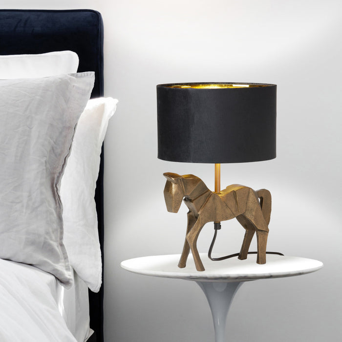 17.75 H Gold Origami Horse Base Table Lamp with Black Drum Shade on a nightstand next to a bed.
