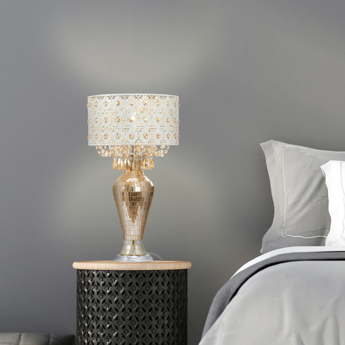 25"H Jeweled Metal and Mosaic Base Table Lamp with Cascading Crystals on a nightstand next to a bed with gray bedding.