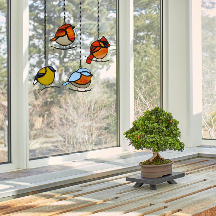 4.5" H Bird Quartet Stained Glass Window Panel Set hangings in front of a large glass door with a potted plant on a wooden table surface.