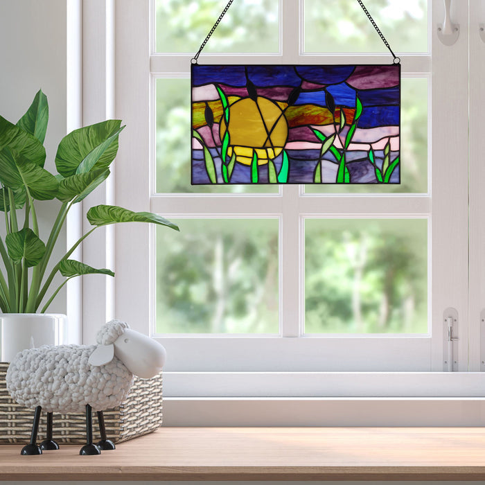 8.25"H Purple Cattails at Sunset Stained Glass Window Panel, suspended in front of a white window. The scene is styled with a potted green plant and a white sheep decoration on a wooden surface.