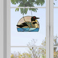 Loon Stained Glass Window Panel hanging in front of a window with a view of trees.