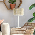 Sasha White Metal and Tan Wood 25-Inch Table Lamp in a living room setting with a wicker chair and plant.